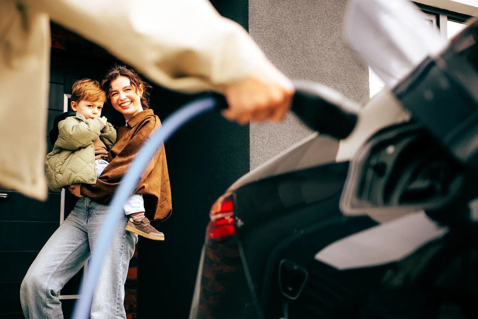 Parent holding a child while plugging a charging cable into their electric car at home